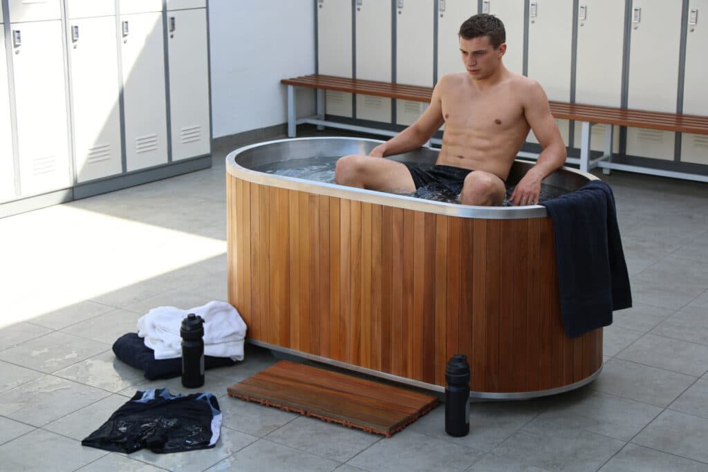 Athlete using wooden cold plunge tub in commercial gym locker room